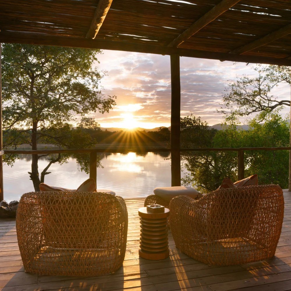 Des fauteuils en osier font face au lever du soleil sur l'eau calme de la lagune à Chindeni Bushcamp