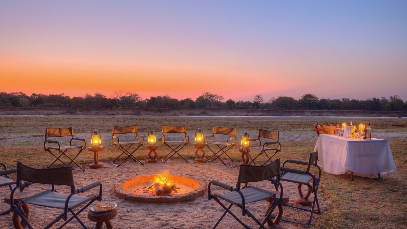 Des chaises pliantes encerclent un foyer tandis que le ciel devient orange à Shawa Luangwa Camp