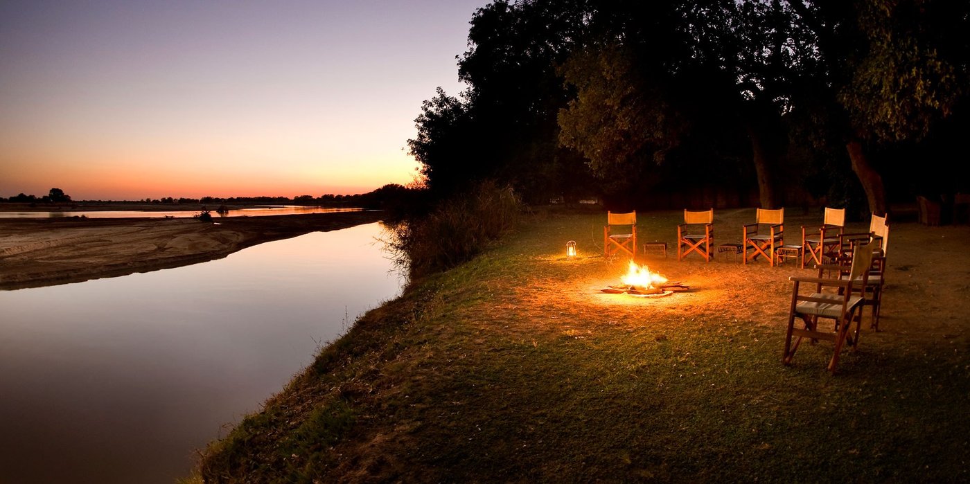 Des chaises de toile encerclent un feu à côté de la rivière au crépuscule, Tafika Camp